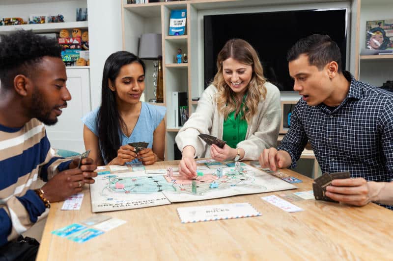 four people playing a board game together, all look excited, while one looks aghast (Photo by Big Potato on Unsplash)