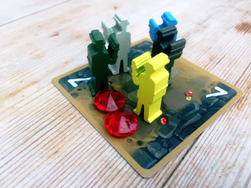 five explorer meeples in different colours and two plastic ruby gems on top of a cave tile