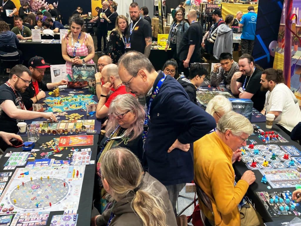 a number of people sitting a tables playing board games while a few are standing and teaching the games at UK Games Expo 2024