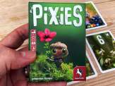 someone holding the Pixies box in their hand above some of the game's cards sitting on a wooden table