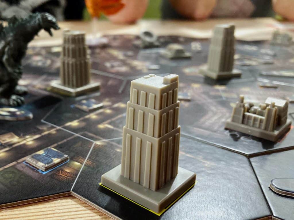 a number of plastic miniature buildings on the modular game board with a Godzilla miniature in the distance